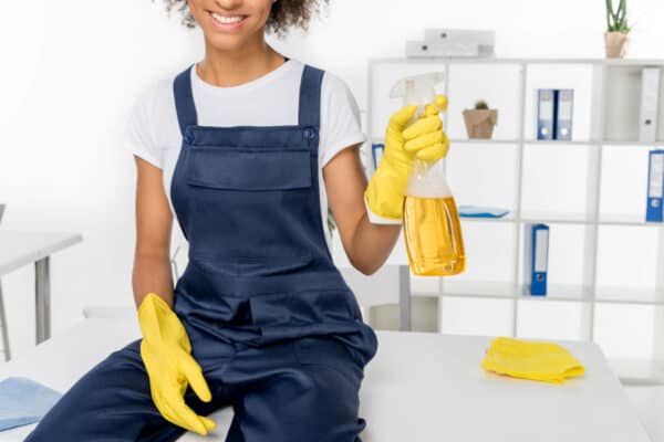 Professional cleaner in Austin, TX wearing yellow rubber gloves and overalls, holding a spray bottle with a cleaning mixture for a water dispenser